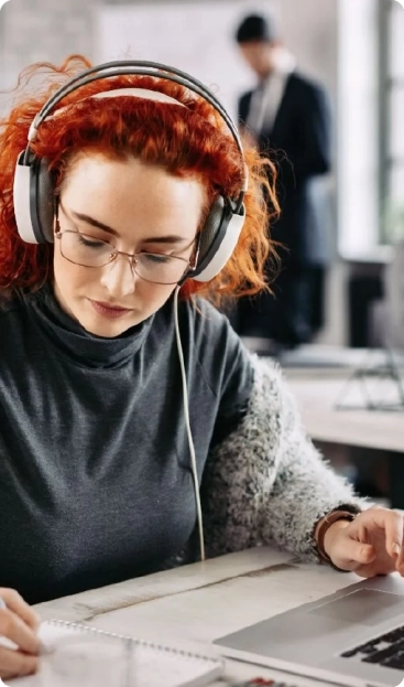 A woman with red hair, wearing headphones, focused on her laptop as a virtual research assistant