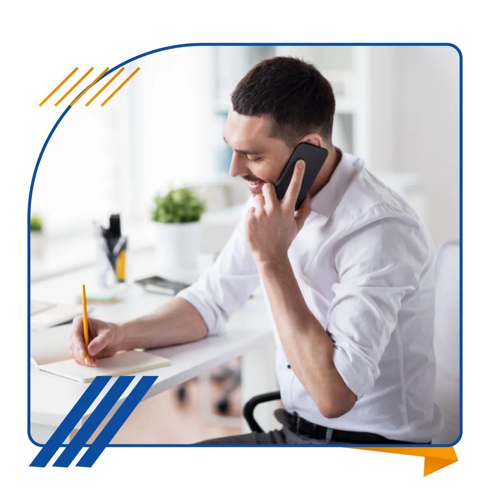 A man engaged in a phone conversation and writing on paper while seated at his desk, focused on his work environment